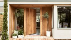 A wooden oak front door opening into a home. Plants in white pots on wither side of the door, red brick porch flooring.