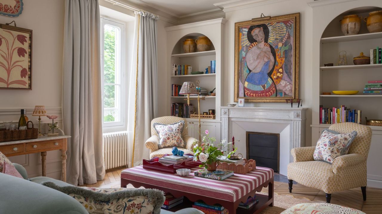 neutral London living room with a fireplace, alcove bookcases, large art, vintage objects, a striped ottoman, and two armchairs facing a blue couch