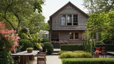 A lush, tiered backyard garden with manicured hedges, stone pathways, and a large outdoor wooden dining table. In the background stands a weathered two-story grey shingle house surrounded by mature trees.