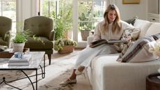 Designer Shea McGee sat on a neutral couch reading a coffee table book, with large patio doors behind her, two green armchairs and a styled marble coffee table on a patterned rug