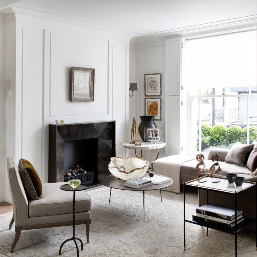 A white living room with neutral furniture, large windows, white panelled walls, a patterned pale Persian-style rug, and a black marble fireplace in the center of the back wall. To the left of the fireplace is a tall shelving unit with books and decorations.