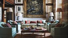 living room of a traditional jacobean home with wooden beams and a large tapestry hung on the wall, styled with artwork, a light green sofa, four tub chairs and a striped upholstered ottoman