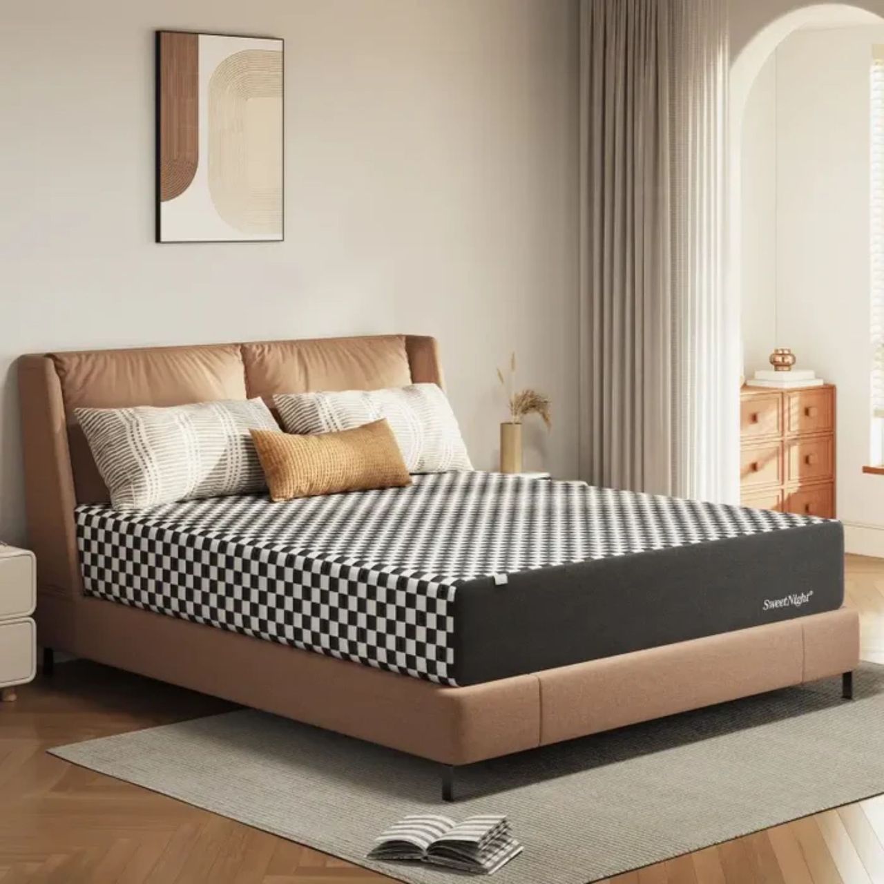 A black and white chequered SweetNight Prime Memory Foam Mattress pictured on a light brown upholstered bed in a cozy bedroom. On top of the bed are two patterned black and white pillows, and a rectangular burnt yellow cushion. Beneath the bed is a large grey area rug, with an open book pictured on it, and the floor is wood. To either side of the bed are modern white bedside tables, and the one on the right also features a gold vase with dried plants in it. Above the bed is a minimalist neutral artwork, and the walls are cream. To the right of the bed is an open curtain, with a smaller room section seen behind with a wooden chest of drawers and small gold vase.