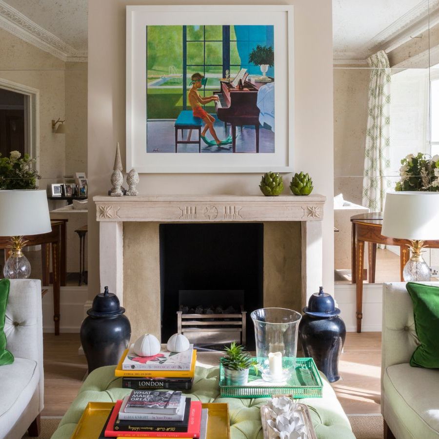 A cream and green living room with a mirrored wall, large mantelpiece with colorful artwork above, two matching cream couches with green cushions and a green coffee table between.