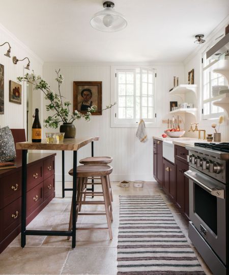A cottage kitchen with white paneled walls, burgundy cabinetry, a striped runner, and open shelving filled with tableware and vintage decor