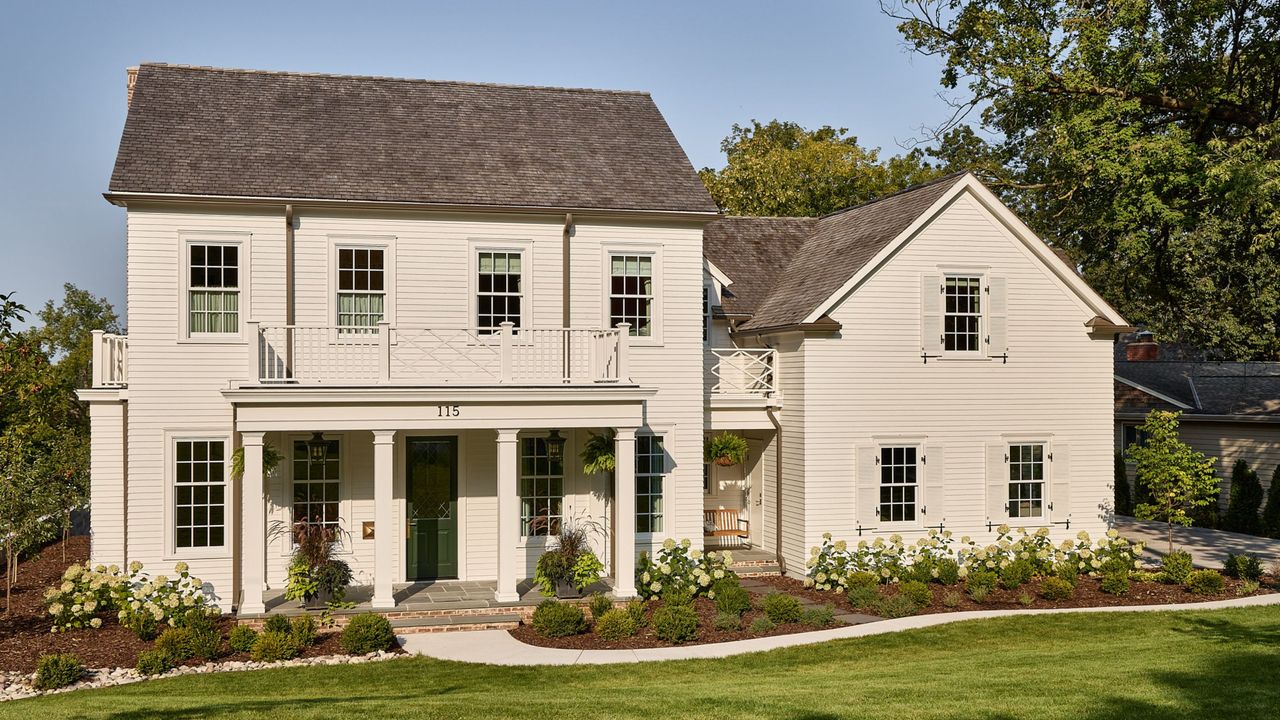 A large home exterior with warm white timber-clad walls, a front porch with four pillars, traditional-style windows and a dark green front door. A garden at the front of the house with lush green grass and a curved path. 