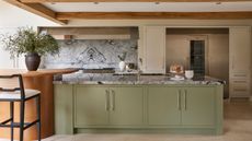 A kitchen with a large green painted kitchen island and marble countertop, built into a large orange kitchen table with black bar stools. A floor-to-ceiling fridge freezer unit built in in the background. 