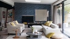 Gray living room, grey graphic patterned wallpaper, white fabric sofa with matching footstool, armchairs upholstered in hexagonal patterned fabric , yellow scatter cushions, grey veneer coffee table, lacquer Boiserie TV panel, lacquer four cube wall unit, sliding patio doors