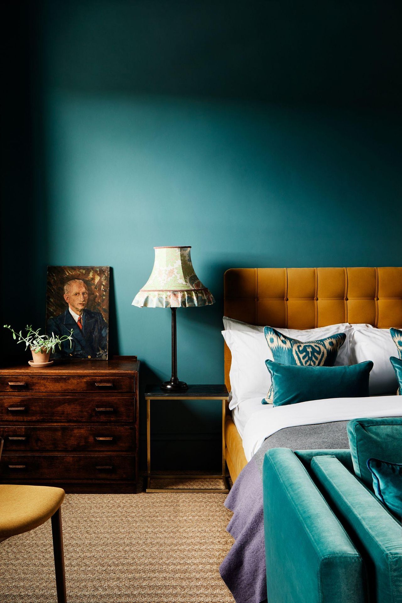 A bedroom with color-drenched teal walls, a mustard bed with white and teal bedding, and a wooden chest of drawers with artwork on it.