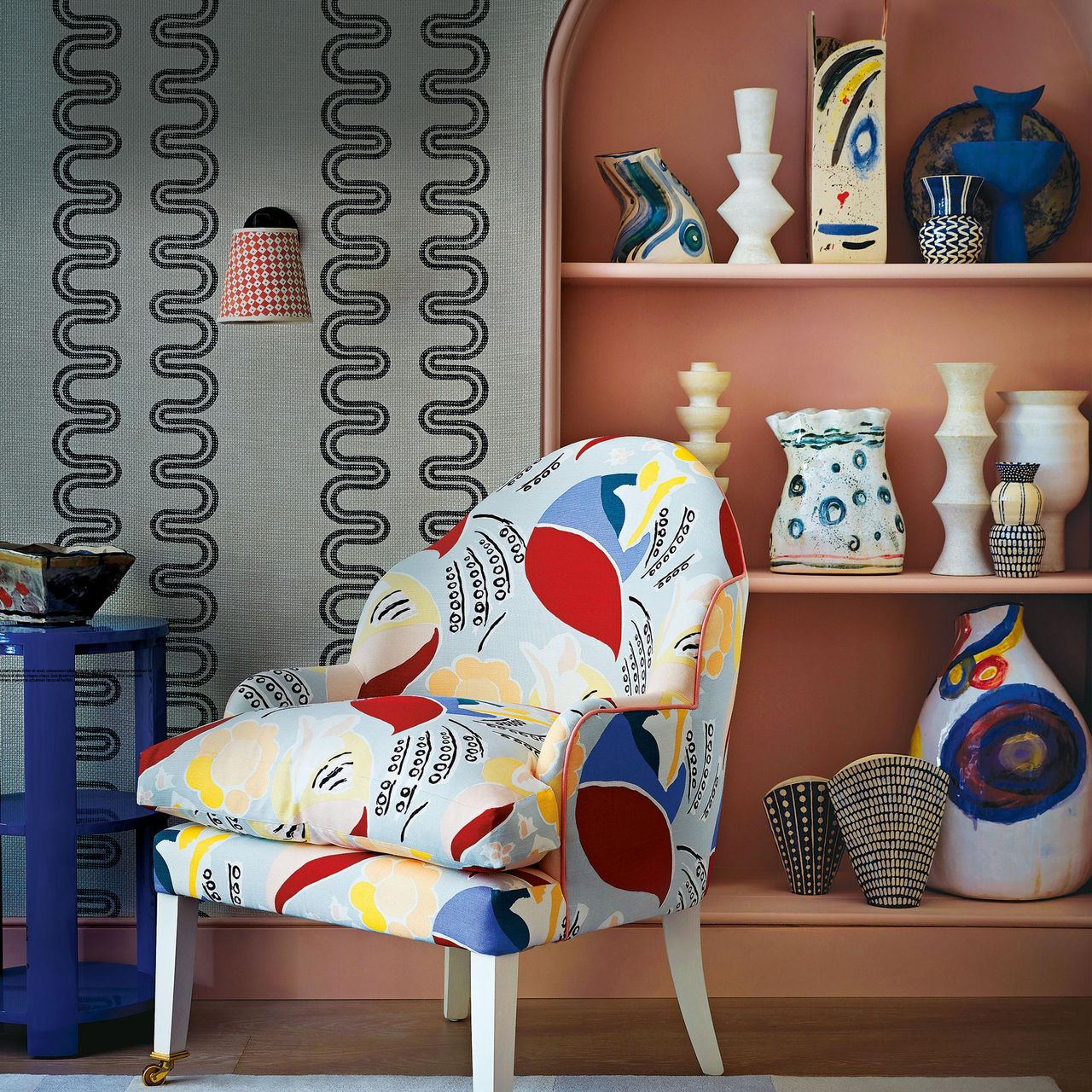 A colourfully covered armchair on a blue rug beside a painted shelf unit of colourful vases.