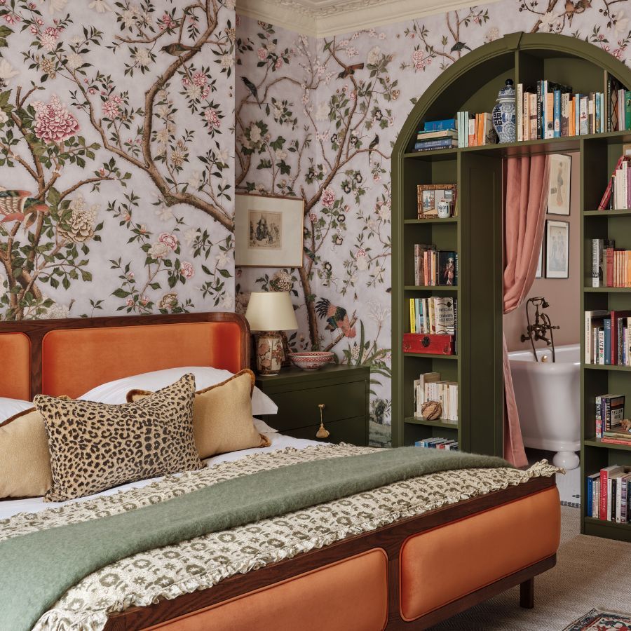 Cozy bedroom with tree and leaf wallpaper, dark khaki green painted door and woodwork, including a built-in arched book shelf storage leading into a bathroom with freestanding tub visible. The bed has a mid century orange velvet header and footer and there are textured pillows and throws on the bed.