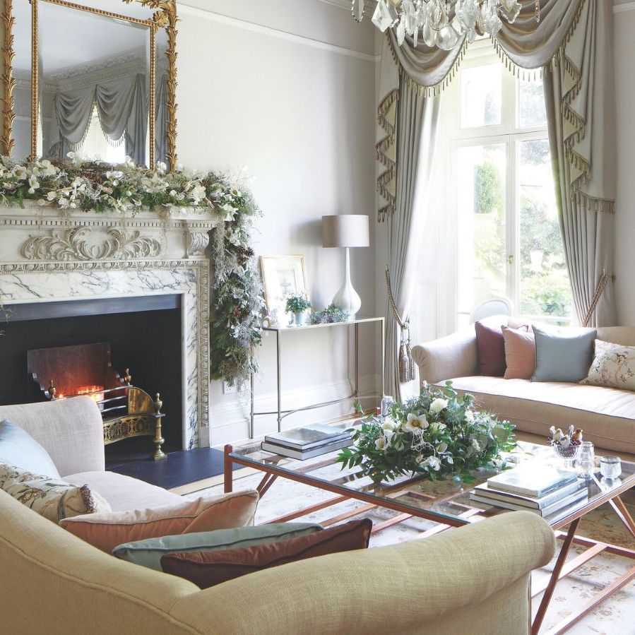 An elegant living room with white walls, white and gold fringe curtains over large windows to the back of the room, and a large marble mantelpiece to the left with a lit fire, large flower garland, and large gold mirror above. In the center of the room is a large glass coffee table with flowers, and upholstered pink and yellow couches and chairs surrounding it with mismatched colored cushions. Beneath the coffee table is a patterned rug.
