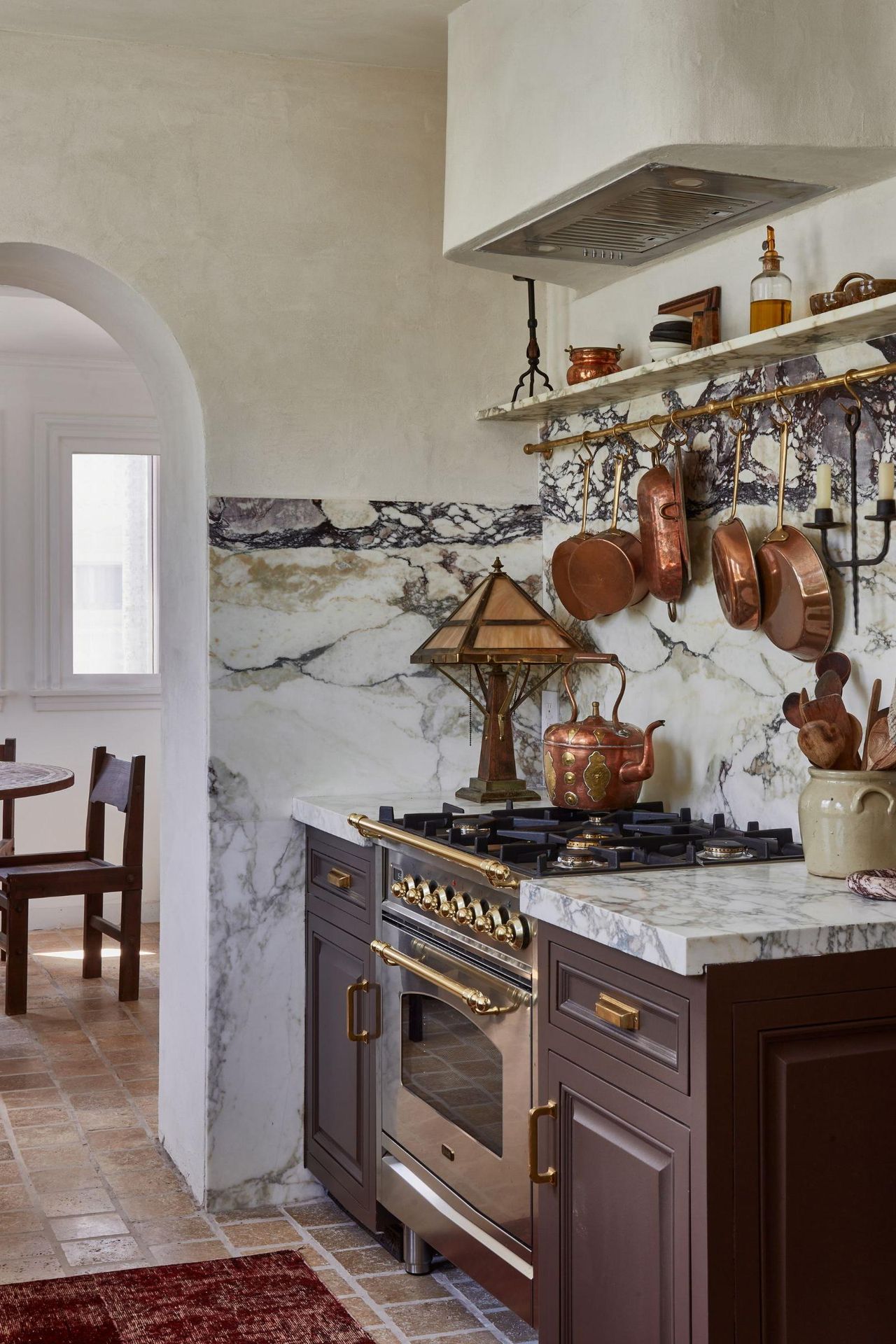 A Vintage kitchen with brown cabinets, a marble countersplash, a brass pot rack on the wall, and a range cooker topped with a vintage copper teapot