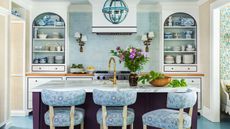 Blue and white kitchen with blue tiled backsplash, built-in alcoves with open shelves of china, aubergine painted island and bar stools upholstered in blue floral fabric