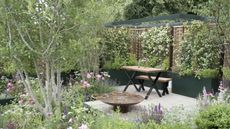 A modern garden with climbers, trees, flower borders, a water feature, and patio tables and chairs at the Hampton Court Flower Show