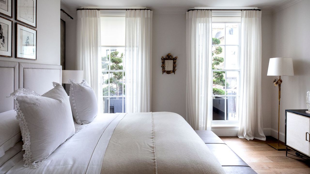 A white bedroom with a double bed with white bedding, white walls, wooden flooring, a gold lamp with white lampshade, black and white cabinetry, and two large windows with sheer white curtains.