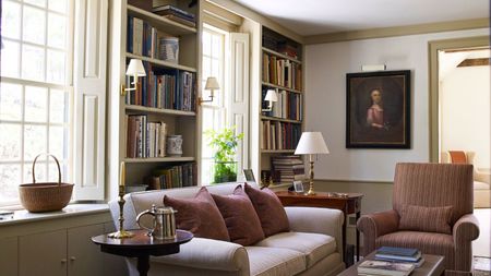 A cozy living room with build in book shelves between windows. A cream sofa in front beside a brown upholstered arm chair. 