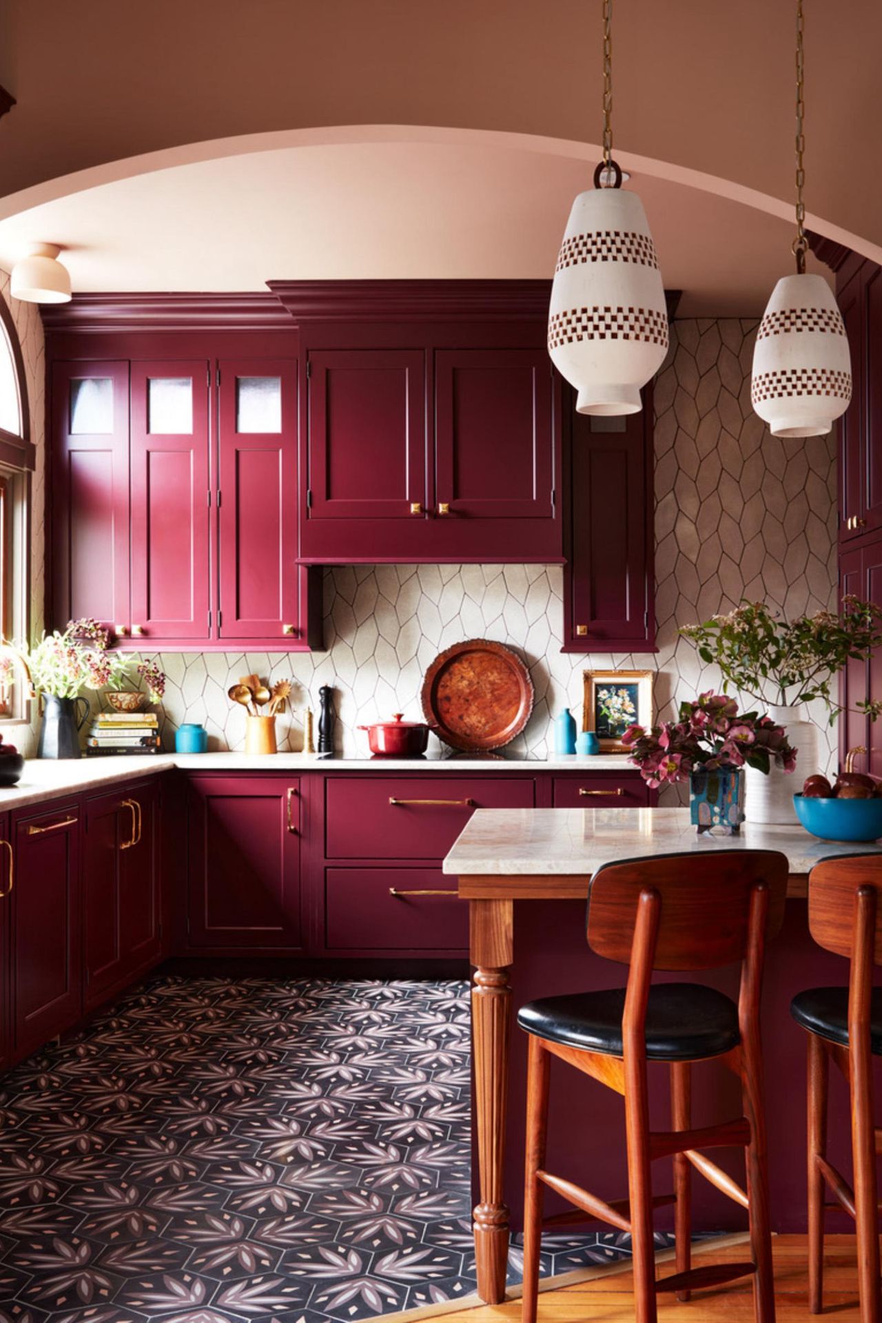 burgundy kitchen with burgundy cabinets, blue patterned floor tiles, kitchen island, pearlescent tiled wall/backsplash, plaster pink walls and ceiling, white pendant lights, brass hardware