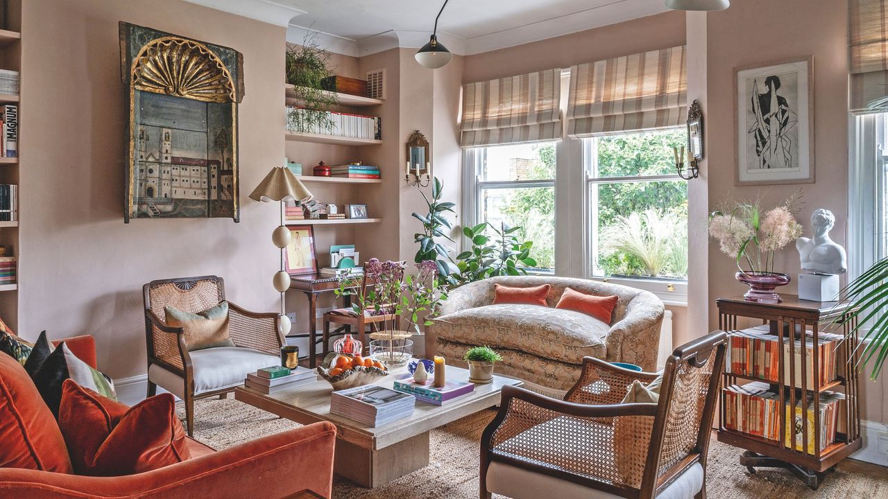 A warm, eclectic living room featuring dusty pink walls, a burnt orange velvet sofa, and woven cane armchairs arranged around a stone coffee table