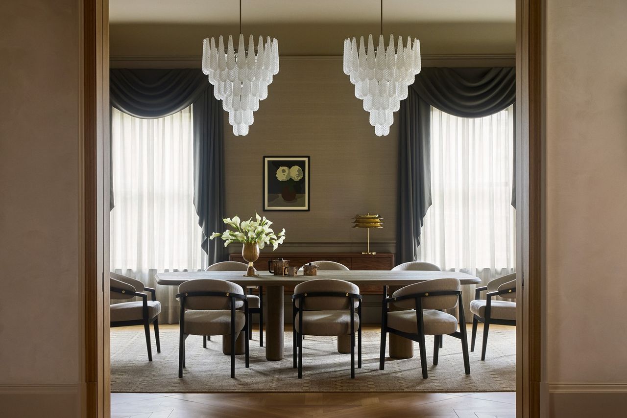 A symmetrical, sophisticated dining room viewed through a doorway. The space features a long wooden table surrounded by curved grey armchairs, two large tiered glass chandeliers, and tall windows with dramatic charcoal grey swags and sheer curtains