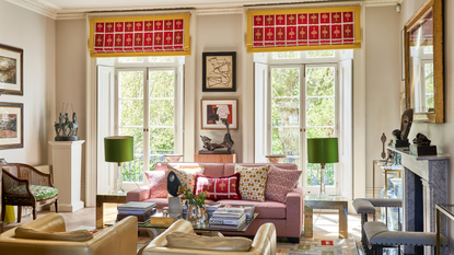 a living room with two yellow accent chairs, a pink couch with lots of pillows, two large windows with red shades, and lots of decor on the coffee table and throughout the room