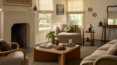 a living room with a large fireplace, a square coffee table, an accent chair with pillows on it, a small side table, and fabric blinds in front of the windows in the corner
