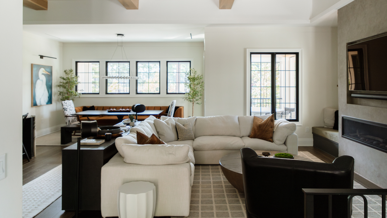 an inviting living room with a large cream sectional, a leather accent chair, a patterned rug, pillows, a dark console table behind the couch holding decor, and a TV built into the wall above a fireplace 