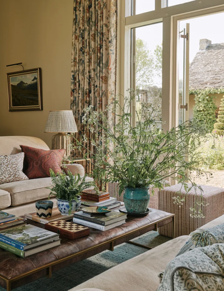 english country decorated living room with soft yellow walls and a leather ottoman decorated with a large foliage display by french doors with floral curtains