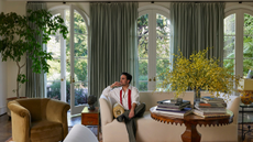 Jake Arnold posing on the cream sofa of a stately sunlit living room featuring a trio of tall arched windows dressed with powdery blue curtains