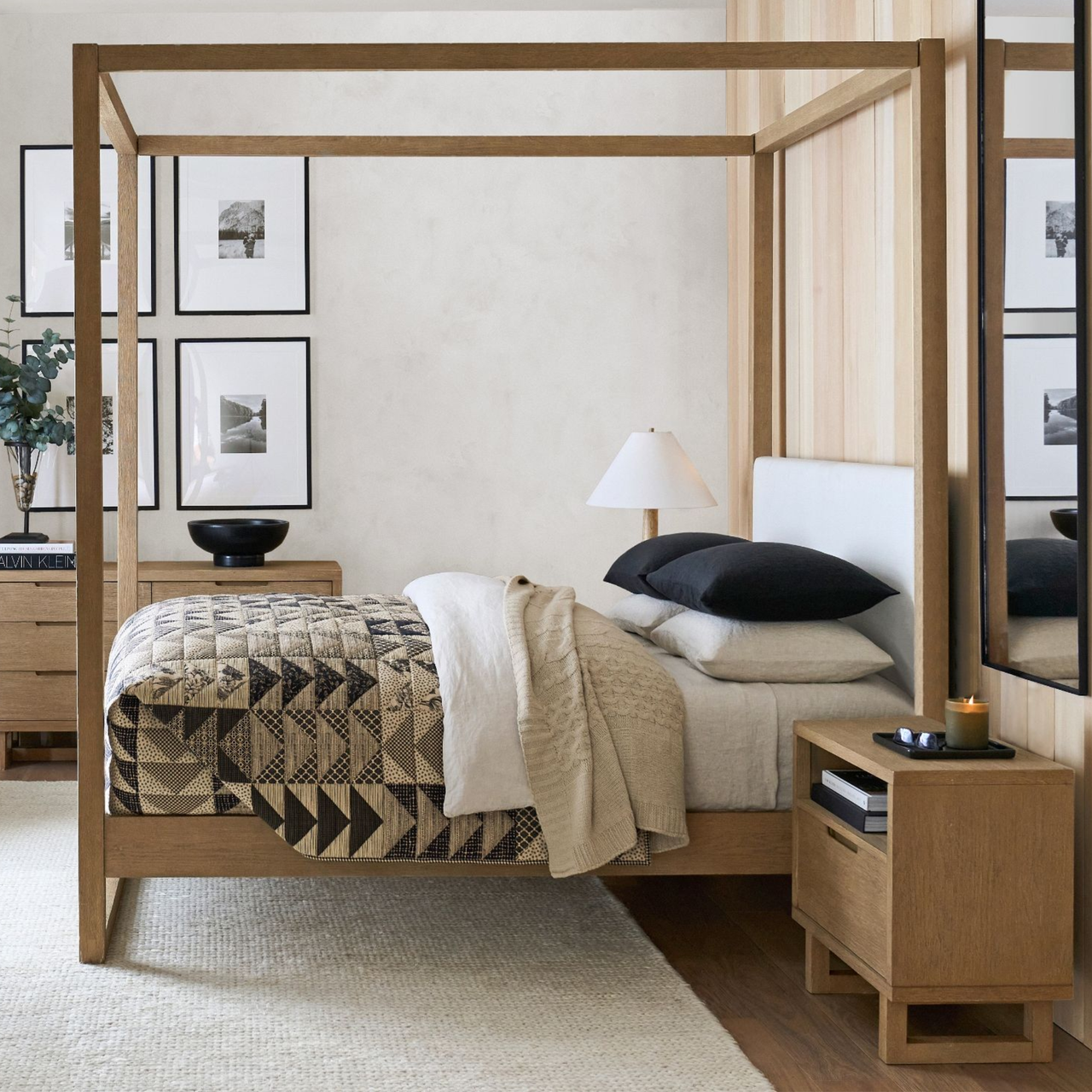 a room with a four poster bed frame, a quilt on the bed, a small side table, and a white rug